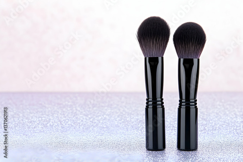 Two black-handled makeup brushes stand upright on a glittery surface against a blurred pink background