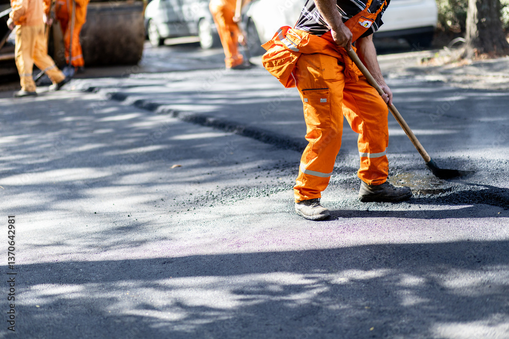 Workers repair road surface with asphalt while wearing bright orange uniforms in an urban setting Workers repair road surface with asphalt while wearing bright orange uniforms in an urban setting