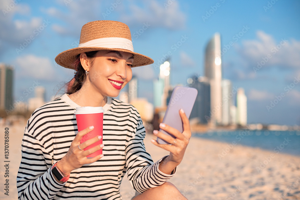 Young woman using mobile phone and holding coffee cup on beach with Abu Dhabi skyline in background