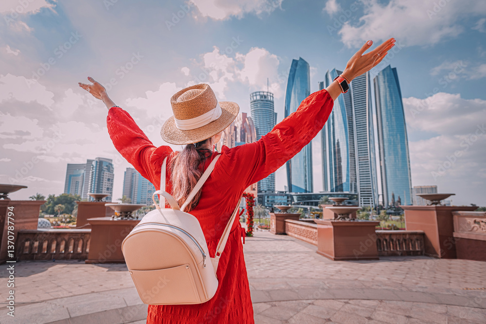 Young woman wearing red dress and straw hat with open arms enjoying the cityscape of Abu Dhabi, United Arab Emirates
