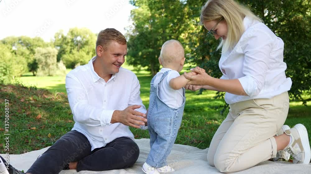 Family is lying on blanket during rest in park. Mom and dad are supporting infant by hand, baby is learning to walk, taking his first steps. Family concept, family rest in nature, first steps of child Family is lying on blanket during rest in park. Mom and dad are supporting infant by hand, baby is learning to walk, taking his first steps. Family concept, family rest in nature, first steps of child
