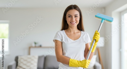 Smiling woman in white t-shirt holding cleaning brush with yellow rubber gloves in bright living room. Professional home cleaning service. Housekeeping and domestic chores concept with copy space.
