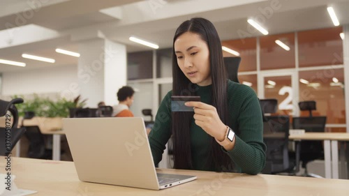 Wallpaper Mural Smiling Asian woman sitting at desk in modern office. Raising fists while holding credit card. Celebrating successful online transaction on laptop. Expressing joy after completing purchase. Torontodigital.ca