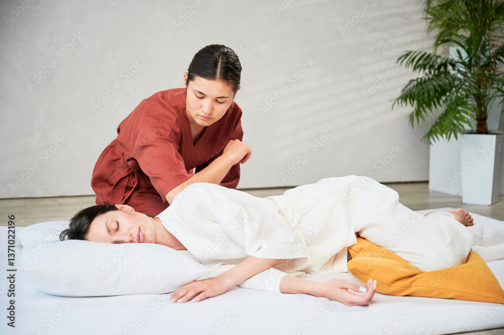 Masseuse making thai yoga massage. Woman therapist doing traditional massage treatment for female patient using elbow. Client lying on mat, meditating, enjoying hand massage in calm atmosphere. Masseuse making thai yoga massage. Woman therapist doing traditional massage treatment for female patient using elbow. Client lying on mat, meditating, enjoying hand massage in calm atmosphere.