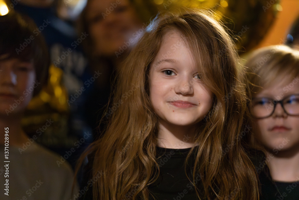 Obraz premium A young girl with long hair smiles at the camera while surrounded by other children at a warmly lit birthday party celebration.
