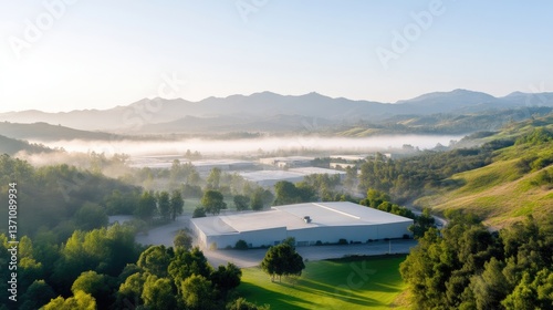 Misty morning view of a large, modern industrial building nestled in a valley surrounded by lush greenery and rolling hills