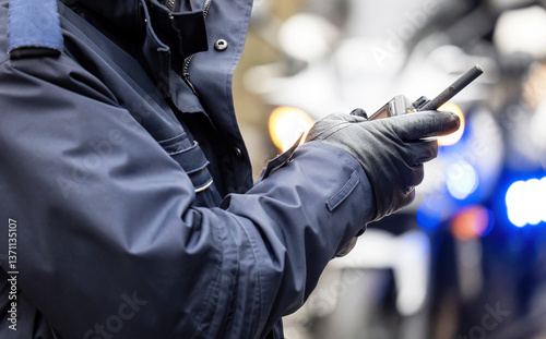 Closeup of a police officer in uniform and gloves holding a two-way radio at emergency scene, blurred crime scene police lights in background, radio communication in public safety services concept