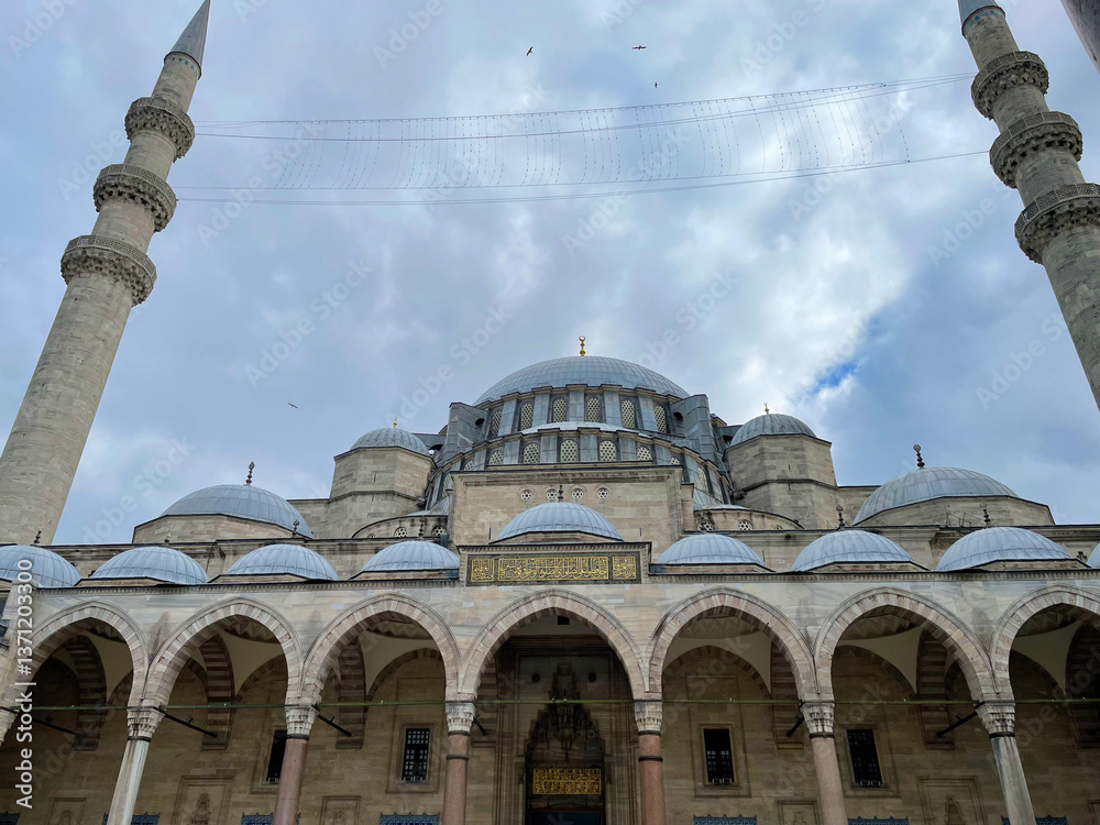 Fototapeta premium Exterior mosque view showcasing bright turquoise dome and twin minarets beneath light cloud cover A vivid turquoise dome and paired minarets rise above the mosque’s stone exterior under cloudy skies