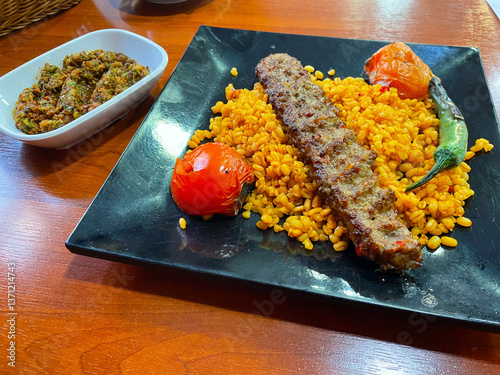 Traditional grilled kebab plate with yellow rice, grilled tomato and dipping sauce bowl Juicy kebab served over yellow rice with grilled tomato and creamy sauce in casual restaurant setting