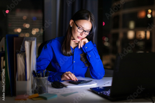 Focused young businesswoman working late with documents, crunching numbers for project deadline, sitting at desk in office