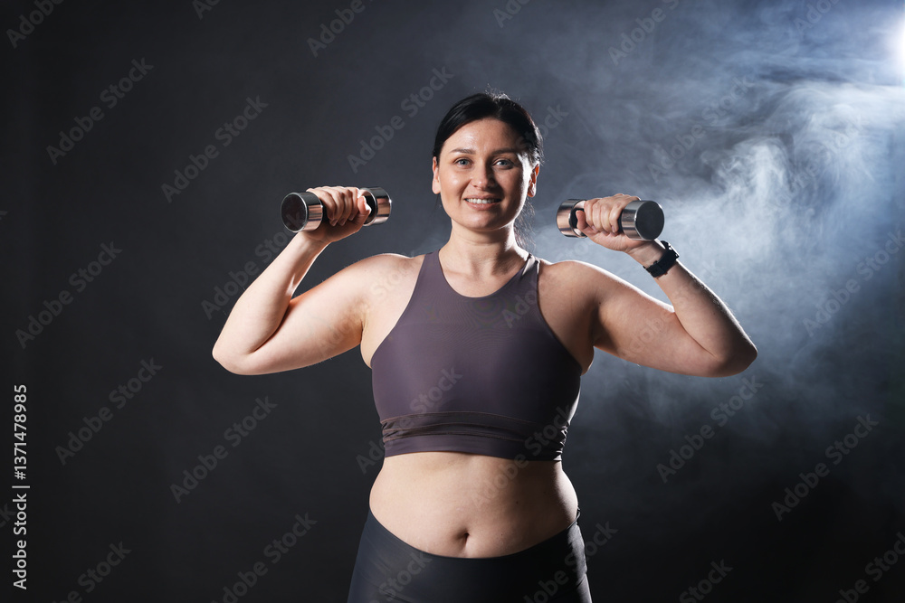 Plus size woman in gym clothes holding dumbbells on dark background with smoke