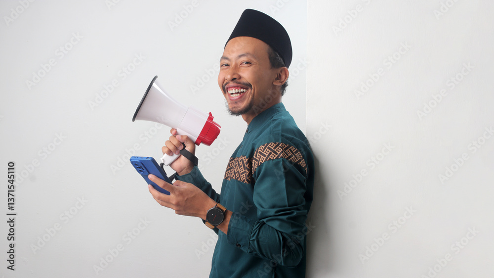 Excited Asian Muslim man leaning against a white wall while holding a megaphone and a smartphone. Isolated on a white background Excited Asian Muslim man leaning against a white wall while holding a megaphone and a smartphone. Isolated on a white background
