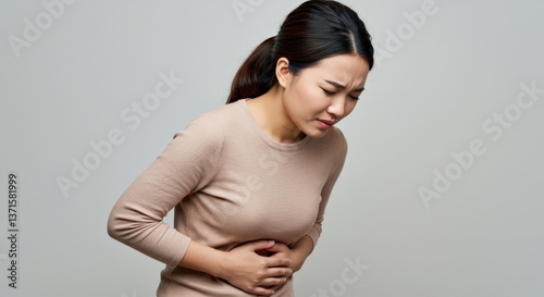 Young Asian Woman Experiencing Abdominal Pain While Standing Against a Neutral Background, Illustrating Symptoms of Discomfort or Illness