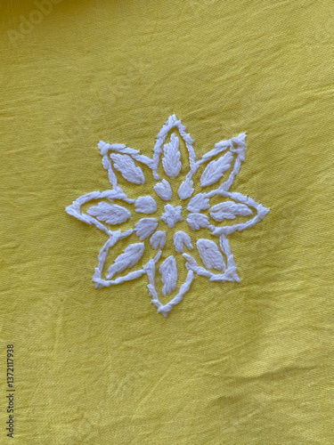 A geometric flower, delicately embroidered with white thread, stands out on lustrous yellow silk fabric.