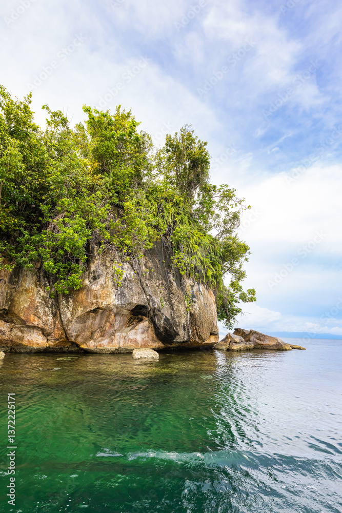 Fototapeta premium Tropical rock island covered with palm trees and dense jungle rises from the calm waters of Triton Bay, West Papua, Indonesia. A stunning seascape surrounded by untouched rainforest and cloudy skies.
