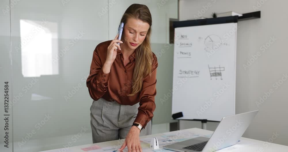 Young professional woman talking on smartphone, coordinating marketing campaign with client or team member by phonecall, reviewing information on laptop while discussing work-related matters by phone