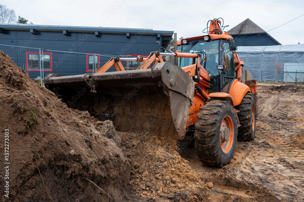 An orange excavator operates on muddy ground with tire tracks, near a modern building with dark walls, red framed windows, and a metal fence. An orange excavator operates on muddy ground with tire tracks, near a modern building with dark walls, red framed windows, and a metal fence.