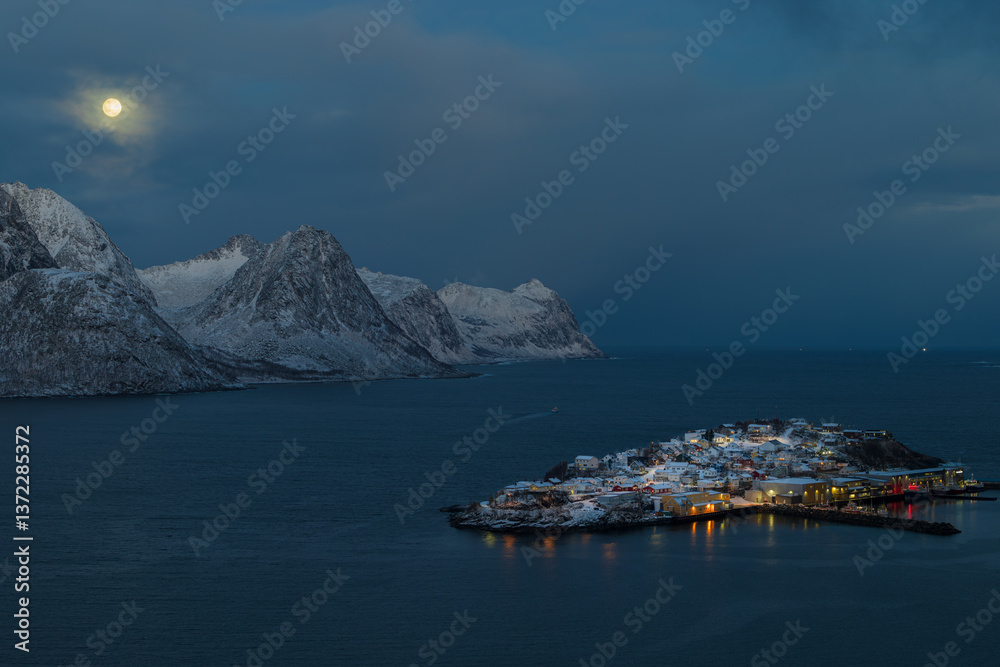 Obraz premium Husøy auf der Insel Senja in Norwegen zur blauen Stunde bei klarer Sicht mit Blick auf leuchtende Häuser im verschneiten Fjord und einem hellen Vollmond über den Bergen am ruhigen Morgenhimmel