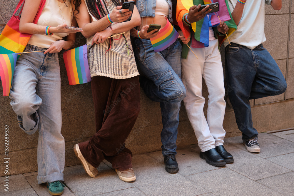 Young people using smartphones and holding rainbow flags are leaning against a wall