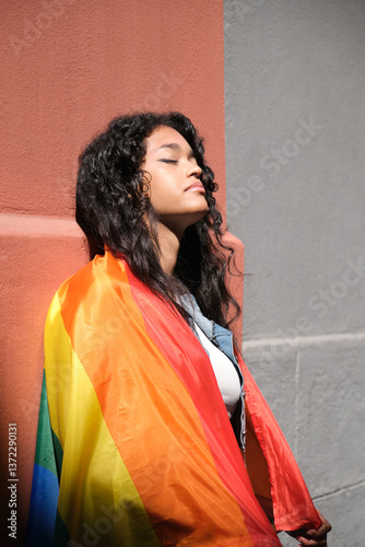Obraz na płótnie Young woman with curly hair wrapped in LGBT rainbow flag enjoying the sun with eyes closed against a wall w salonie