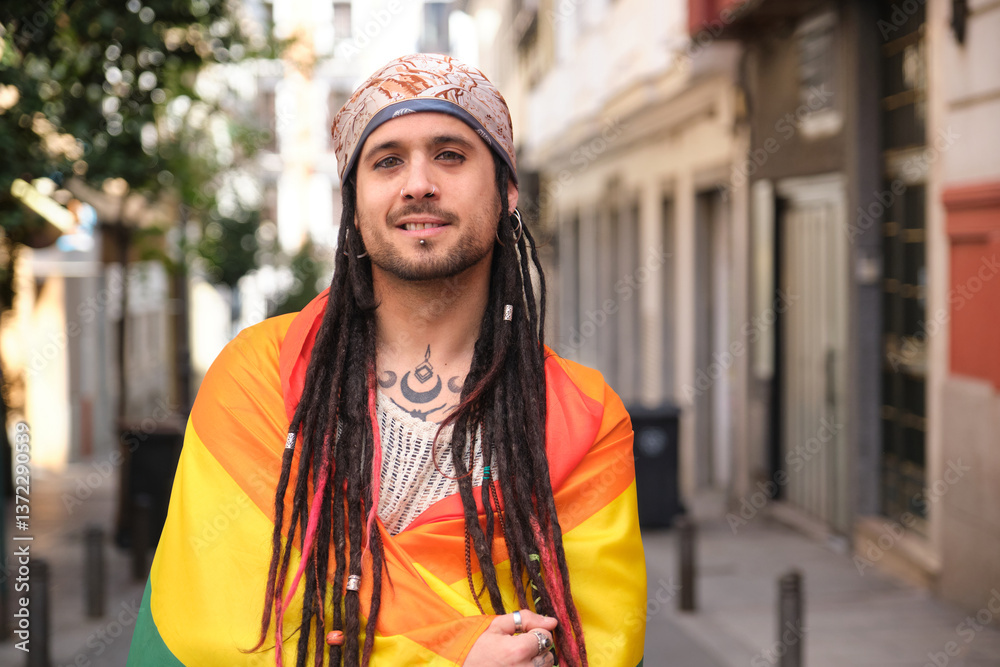 Portrait of a young smiling man wrapped in rainbow flag, symbol of LGBT community, standing in a city street