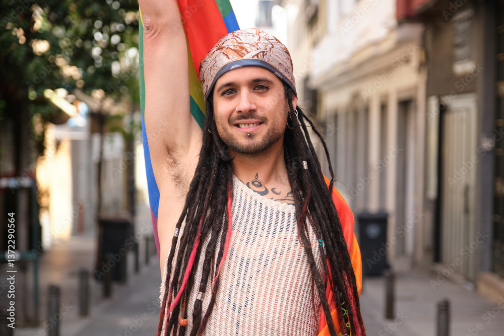 Young smiling man with dreadlocks and bandana raising LGBT rainbow flag while walking in the street