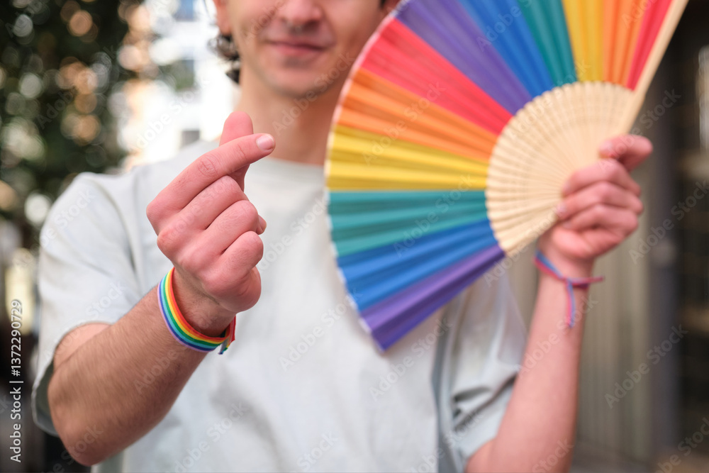 Young man making Korean finger heart and showing rainbow fan celebrating LGBT pride