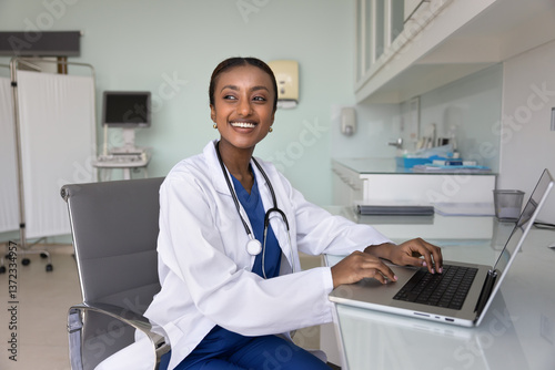 Cheerful young African American medical therapist working at laptop in hospital examination room, looking away with toothy smile, posing for portrait at clinic office workplace
