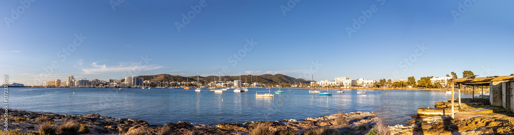 Naklejka premium Sant Antoni de Portmany is a vibrant harbour with yachts and pleasure boats anchored in blue waters. The famous beach and promenade are lined with palm trees and white buildings. Ibiza, Spain