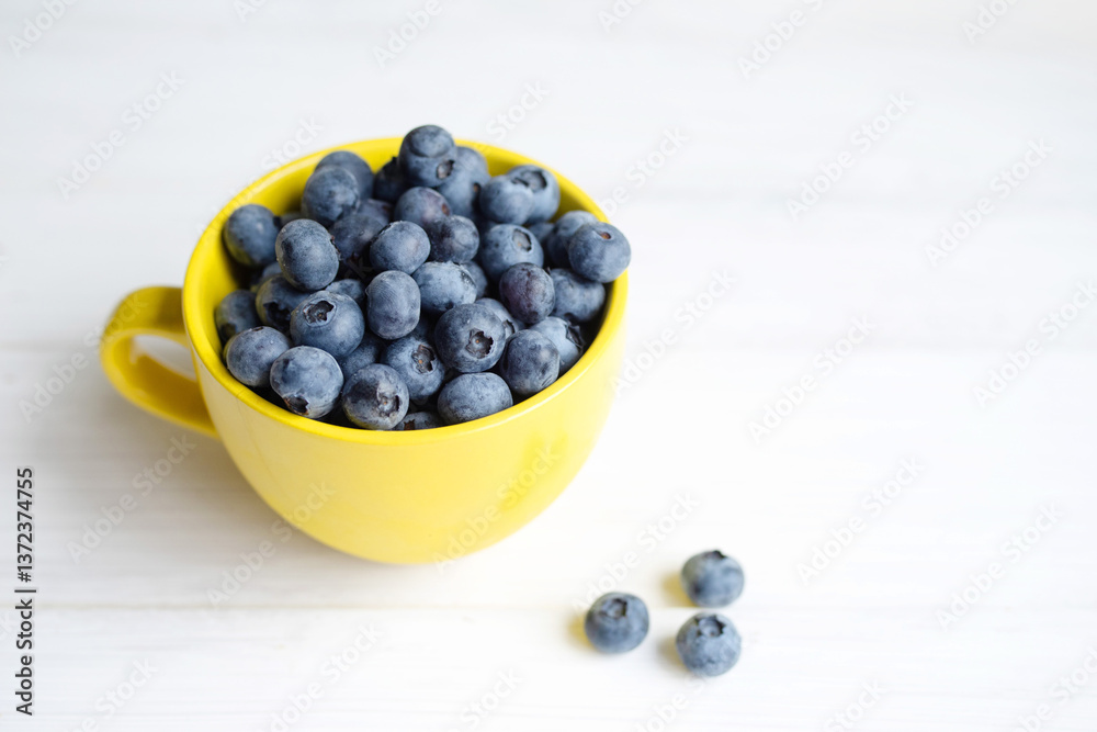 Fresh juicy wild blueberry in yellow mug close-up on white table surface, top view, soft focus
