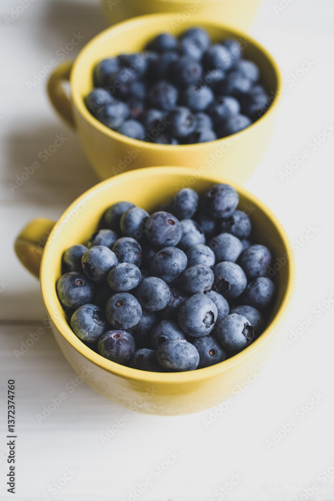 Fresh juicy wild blueberries in yellow mugs close-up on white table surface, top view, soft focus Fresh juicy wild blueberries in yellow mugs close-up on white table surface, top view, soft focus
