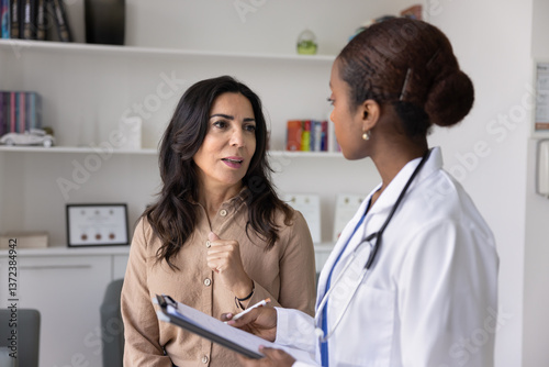 Mature Caucasian woman visiting young African female doctor for consultation, telling about healthcare, sickness symptoms. Physician filling medical form, listening to patient in clinic office
