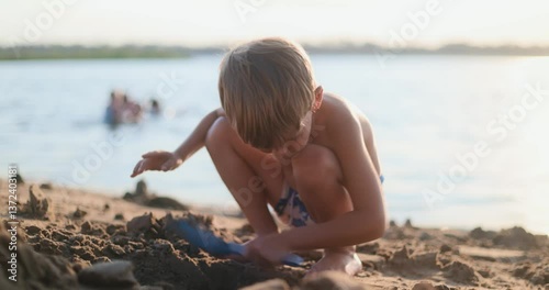 Boy in swim trunks digging sand with blue plastic shovel on beach near water