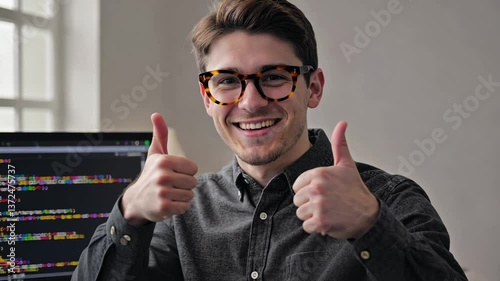 Programmer in glasses sits at a table with a monitor displaying code. Shows thumbs up, as a sign of satisfaction in solving problems and creating innovative solutions in the technology industry.