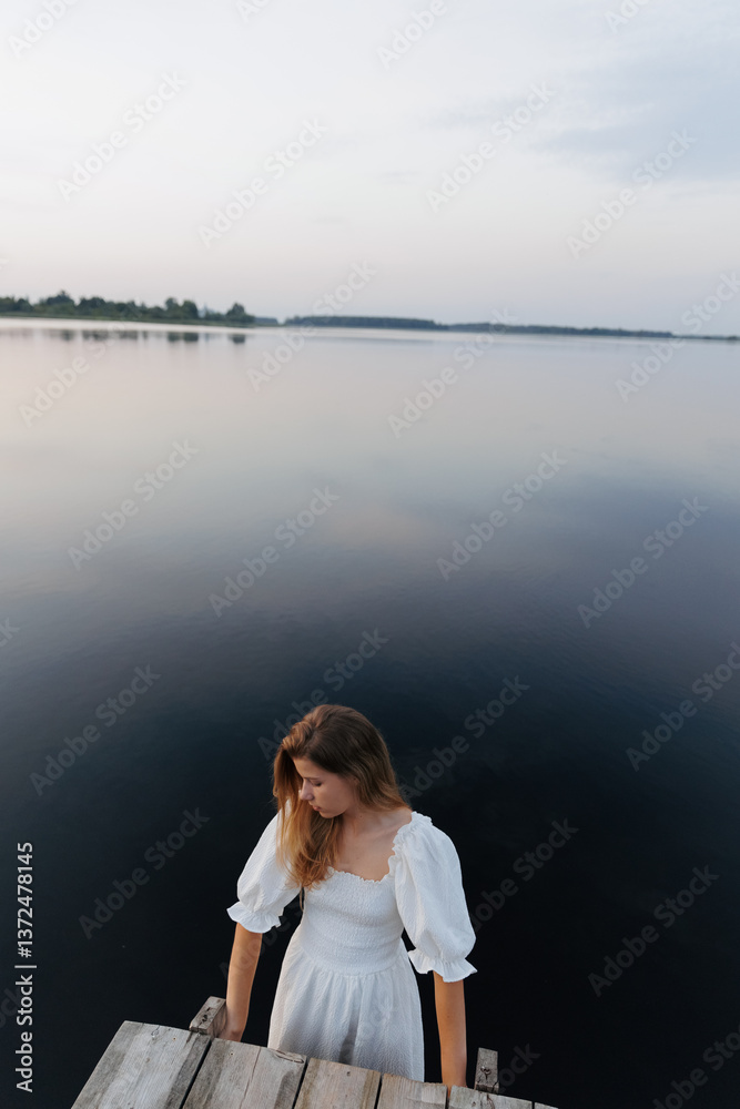 Fototapeta premium A woman in a white dress stands in the water near a wooden pier, holding onto the edge. The evening sky reflects in the calm lake. In the distance, a boat with people adds to the sense of solitude