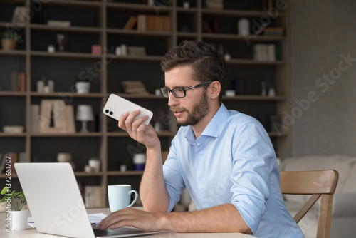 Busy gen z male student freelancer sit by notebook at home office look at screen search for information online dictate thoughts on phone voice recording app use ai assistant make call via loudspeaker.