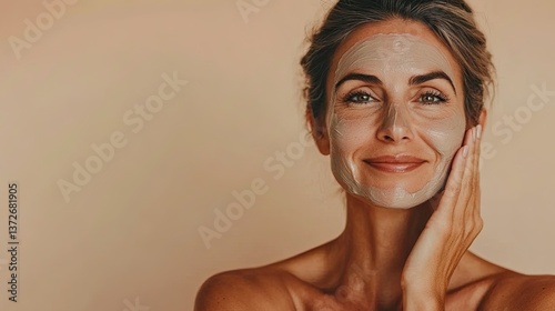 Headshot of lovely middle aged woman applies clay mask on face looks with satisfied expression at camera keeps hands crossed over body has healthy glowing skin isolated on beige studio wall.