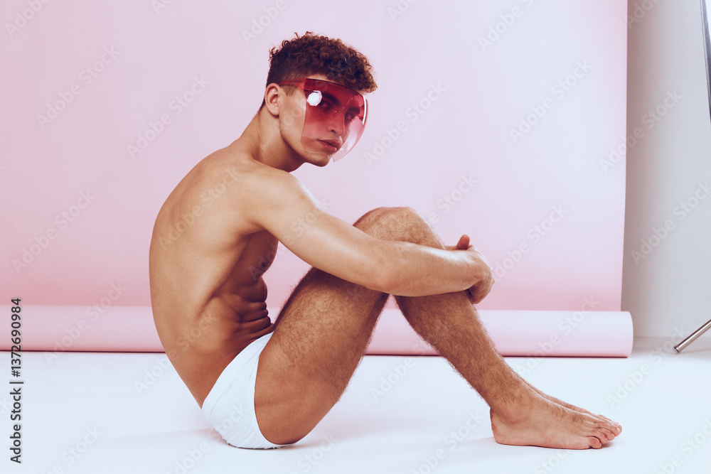 Obraz premium Confident young man in sunglasses sitting on the floor, posed against a soft pink background. Fashion and beauty concept with a modern aesthetic.