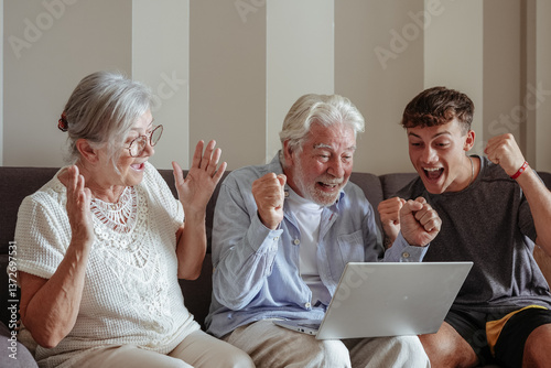Obraz na płótnie Happy multigenerational family of grandparents and young grandson cheering while watching game on laptop sitting together on sofa at home. Senior couple learning how to use computer and technology w salonie