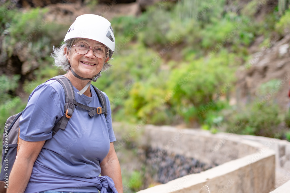 Smiling senior hiker woman in protective helmet sitting to rest during an hike on top of a mountain, elderly lady enjoying trekking day and healthy lifestyle in nature