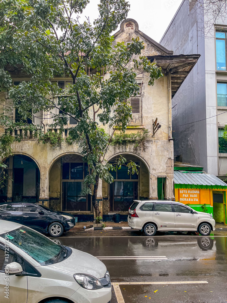Old weathered two-story building with arched openings stands beside a modern structure on a wet Jakarta street, blending historic charm with urban life and parked cars.