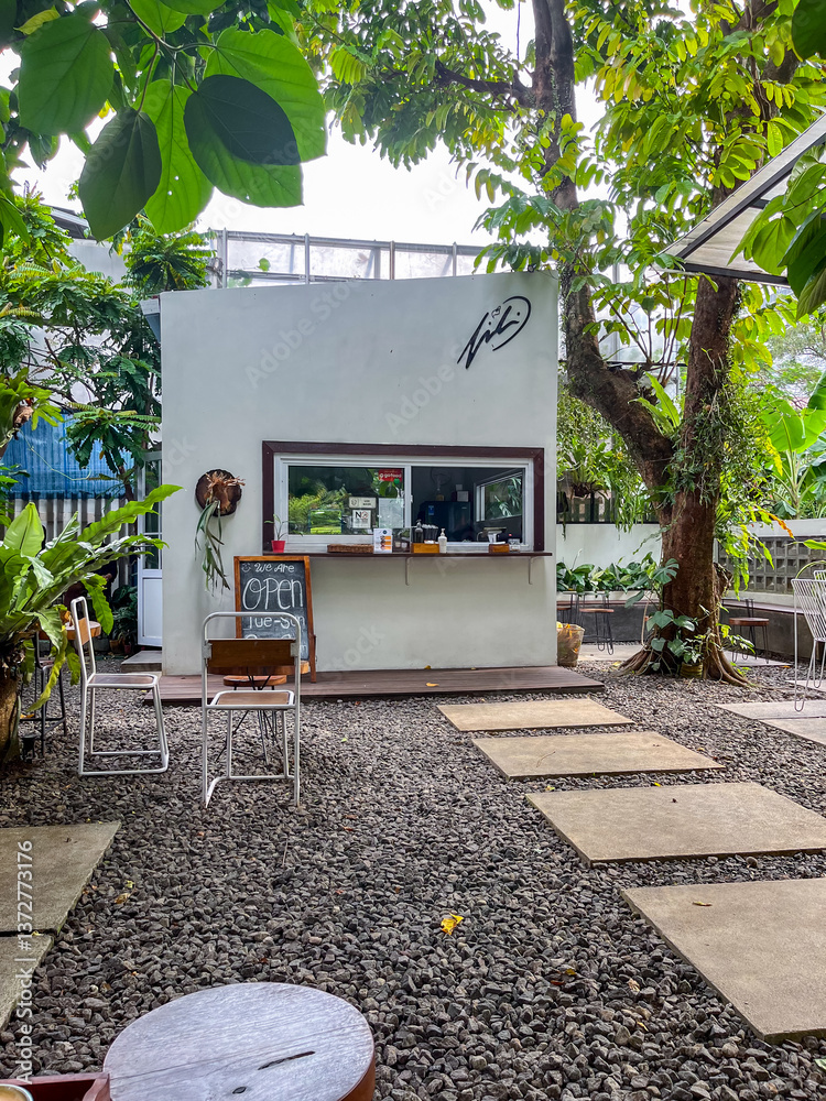 Outdoor café features a small white building with a service window, surrounded by lush trees and greenery. Stone pavers lead to cozy seating areas, creating a relaxed and natural atmosphere.