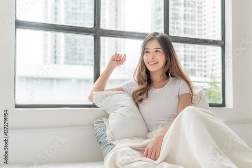 Free Download A young Asian woman with long brown hair, wearing a white t-shirt and wrapped in a white blanket, sits on a bed by a large window, enjoying a peaceful morning at home with soft sunlight streaming in - GFXLoad