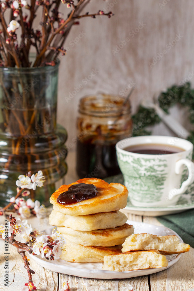 Lenten yeast pancakes with plum jam and tea, and a bouquet of flowering apricot branches on a light wooden table.