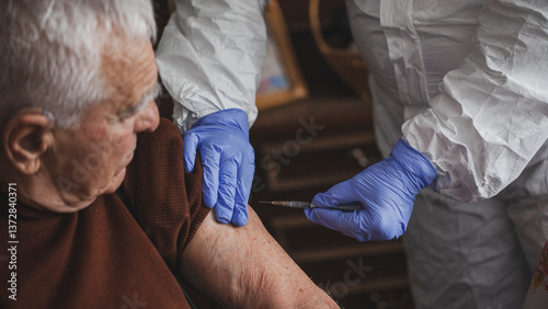 Doctor in protective suit vaccinating elderly man at home during epidemic