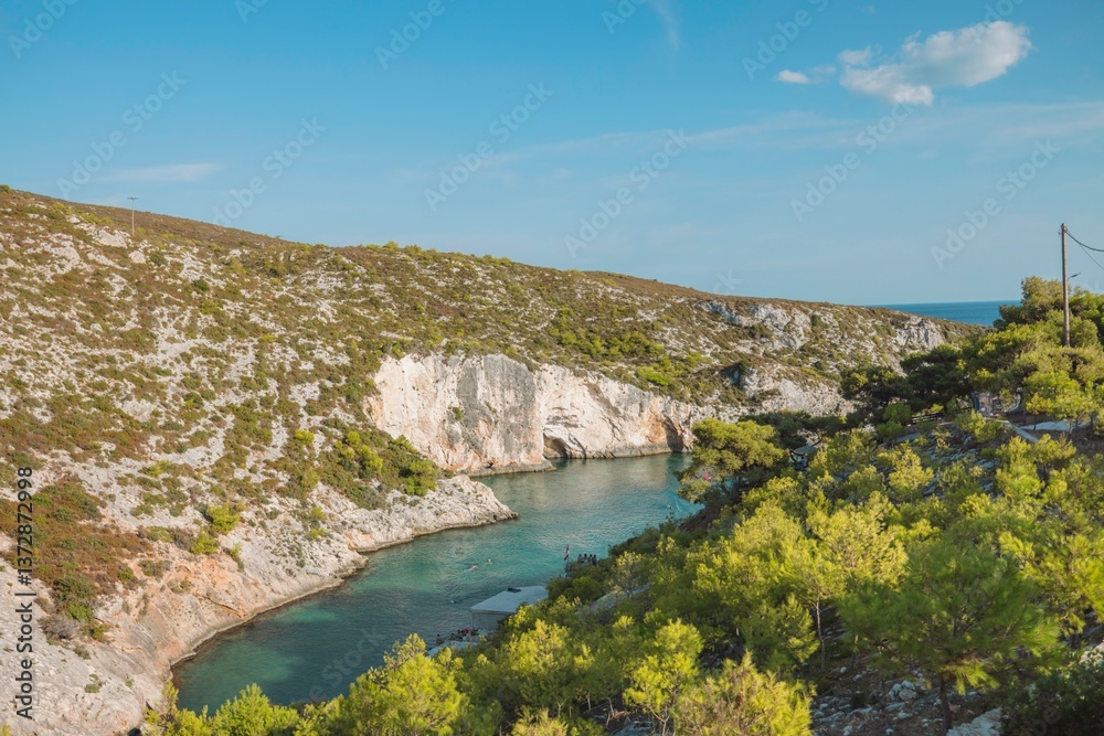 Fototapeta premium Turquoise waters embrace a hidden cove in Greece, surrounded by lush greenery and rocky cliffs. Sunlight creates a tranquil atmosphere perfect for relaxation and exploration