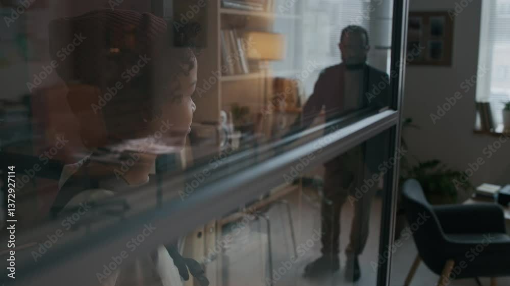 Side shot of little troublesome boy in beanie waiting behind glass partition wall, scared to enter office of school psychologist whose reflection seen on glass Side shot of little troublesome boy in beanie waiting behind glass partition wall, scared to enter office of school psychologist whose reflection seen on glass