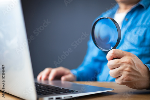 A man in a blue shirt holds a magnifying glass while using a laptop, symbolizing online research, investigation, cybersecurity, data analysis, or scrutiny in digital environments on a wooden desk.