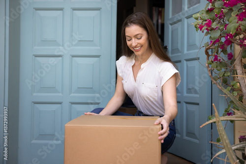 Photography Happy woman sat down to pick up parcel box left by courier on porch, smiling, enjoy convenience of modern doorstep ordered package delivery services