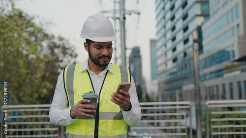 Portrait of male taskmaster in safety workwear holding smartphone and coffee cup on bridge. Man contractor communicates in online chat via mobile phone at construction site